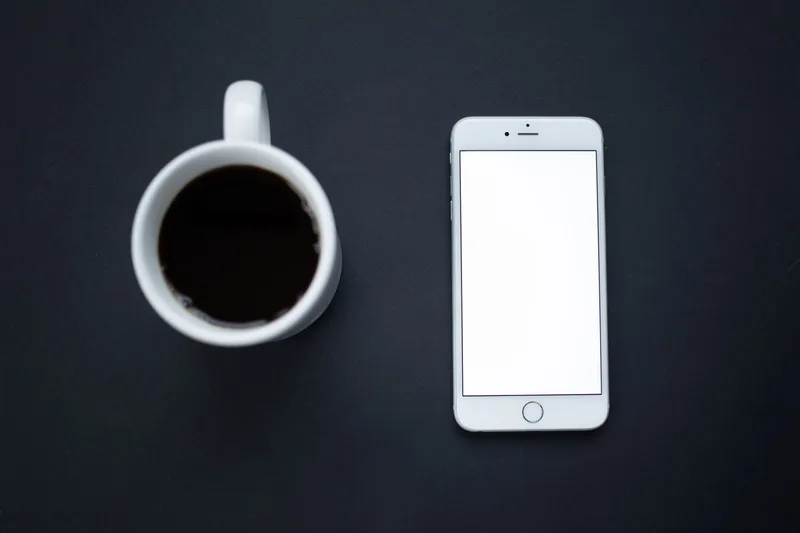 A smartphone with a blank white screen next to a cup of black coffee on a dark desk, representing the minimal starting setup for a faceless Instagram account