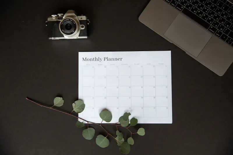 A monthly planner sheet on a desk beside a vintage camera and a laptop keyboard, representing the planning workload behind a consistent posting schedule