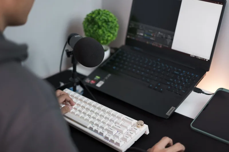 Person at a home recording setup with professional USB microphone, laptop, and keyboard on a clean dark desk