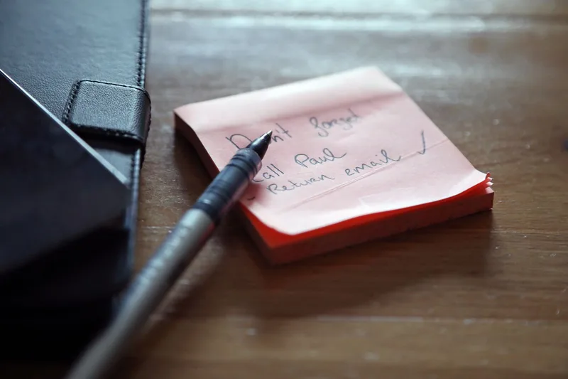 Sticky note with a short handwritten to-do list next to a pen on a wooden desk